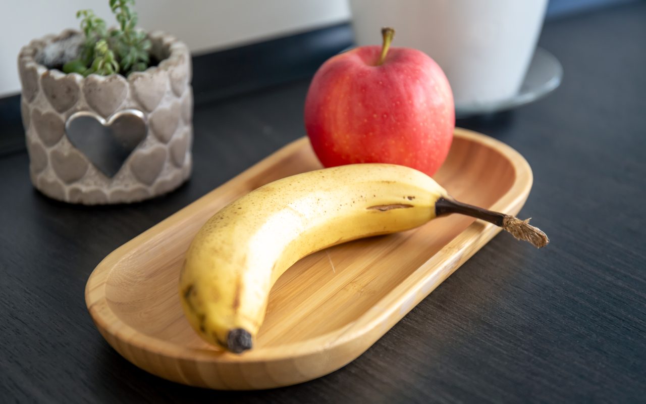 A ripe banana and a red apple on a bamboo tray on a dark kitchen counter