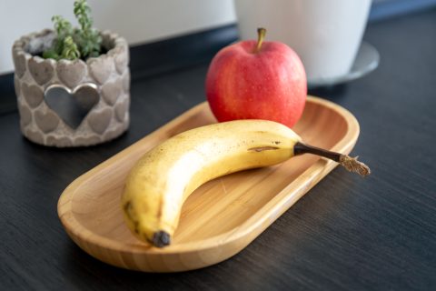 A ripe banana and a red apple on a bamboo tray on a dark kitchen counter