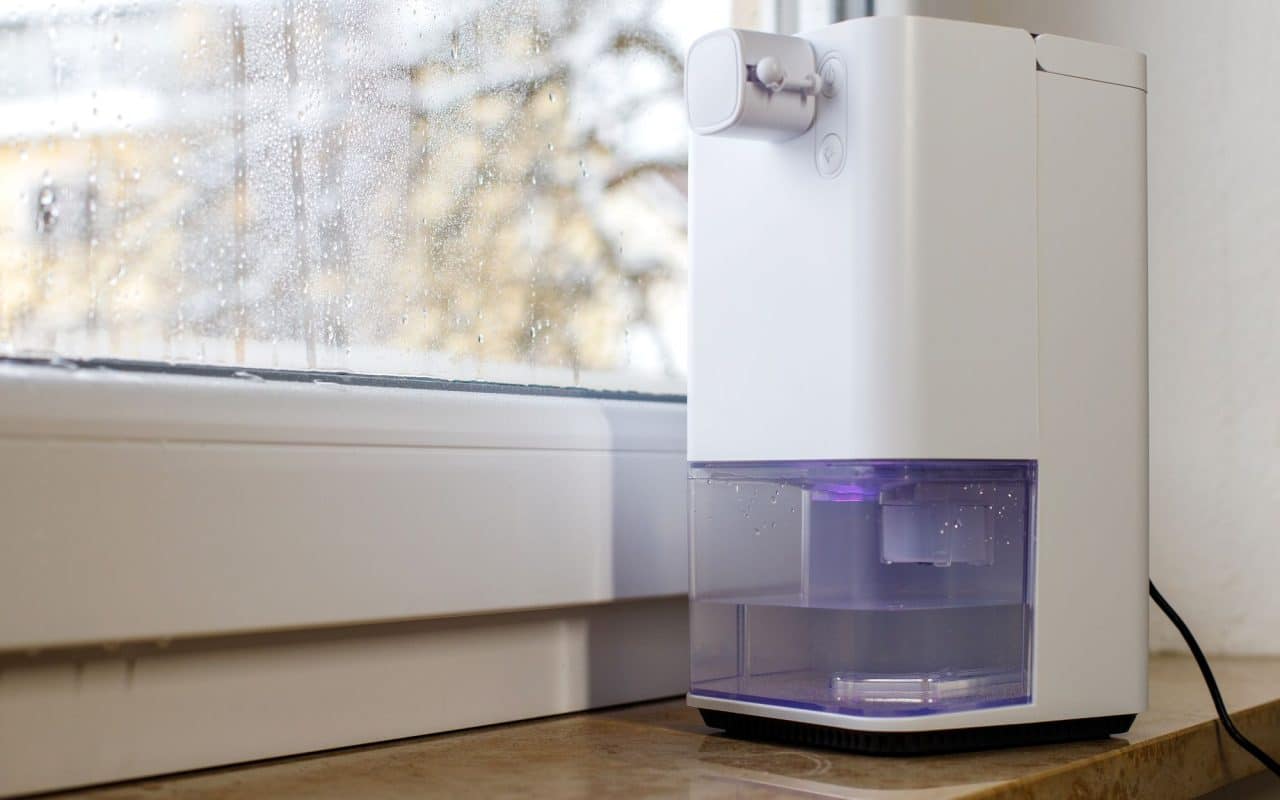Portable dehumidifier running in a damp room beside a window covered in condensation