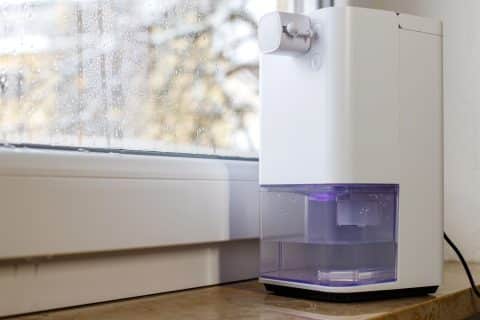 Portable dehumidifier running in a damp room beside a window covered in condensation