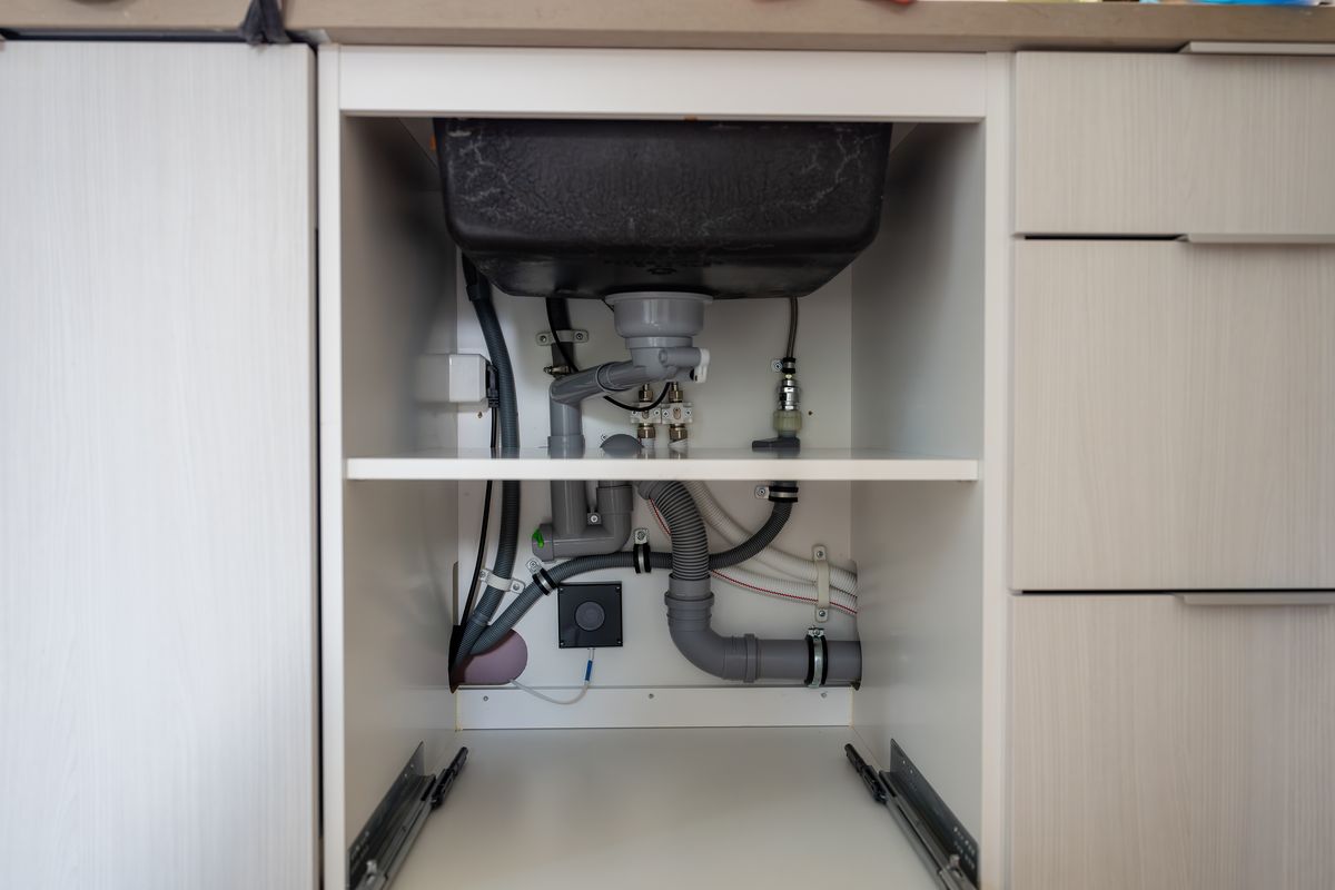 Open cabinet under a kitchen sink showing the drain trap and supply pipes