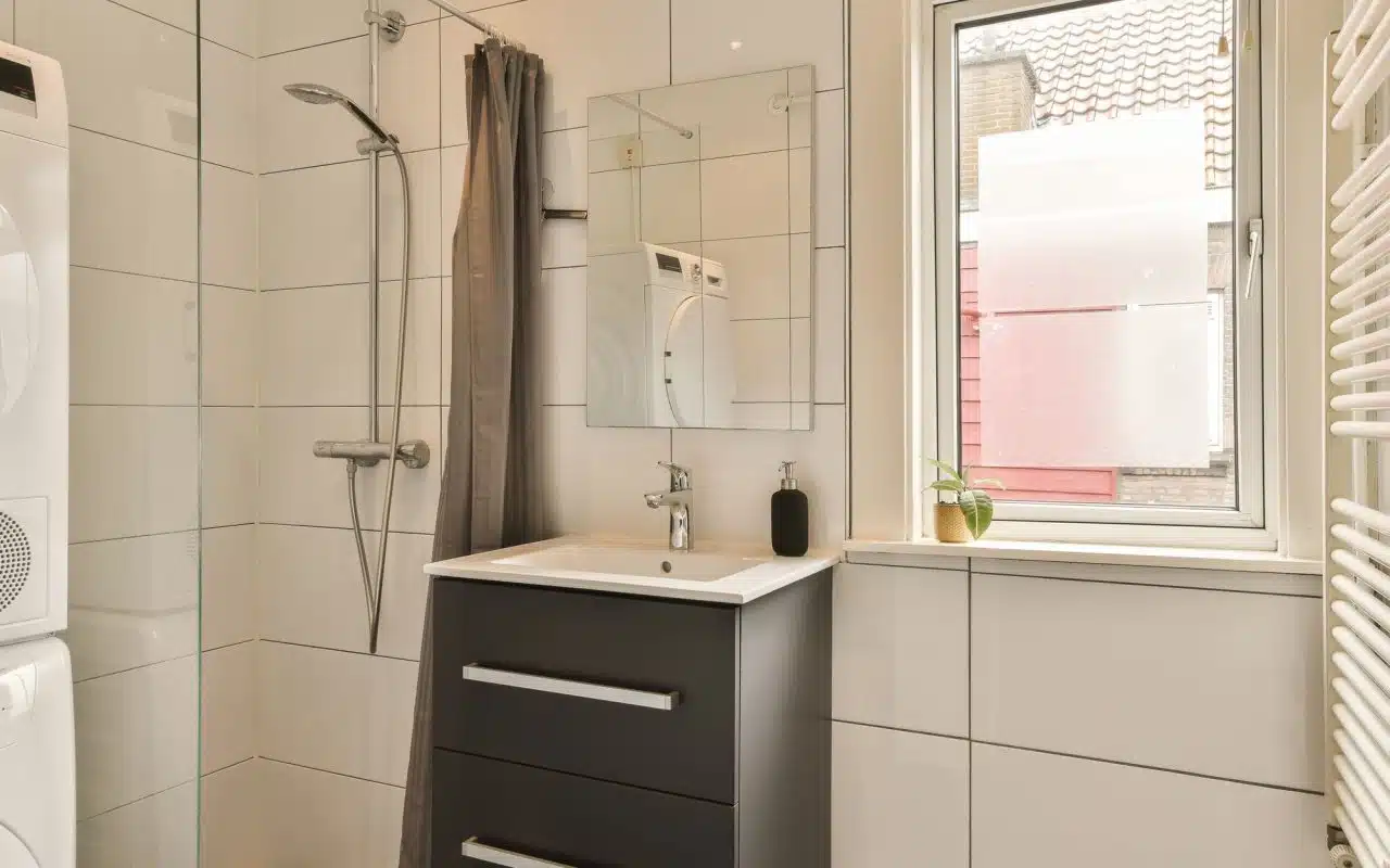 Small bathroom with sink, toilet and corner shower beside a daylit window