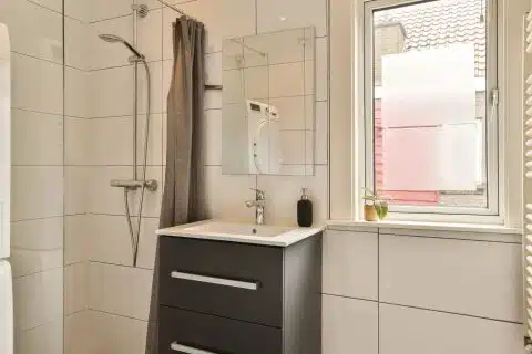 Small bathroom with sink, toilet and corner shower beside a daylit window