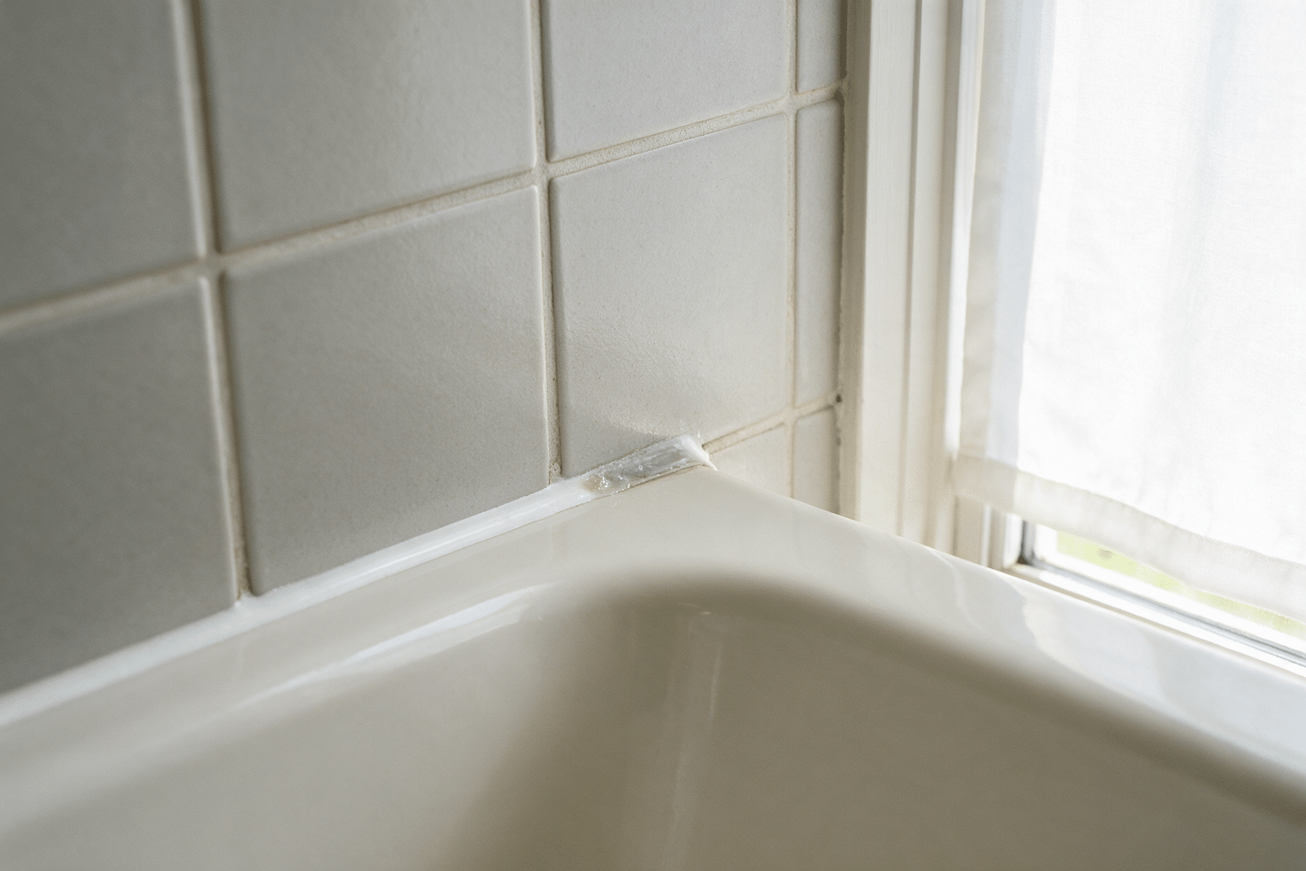 Close-up of a bathtub corner where glazed tile meets the tub rim, with a section of silicone caulk along the joint